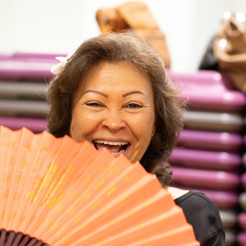 An older woman smiles at the camera while holding an orange paper fan.