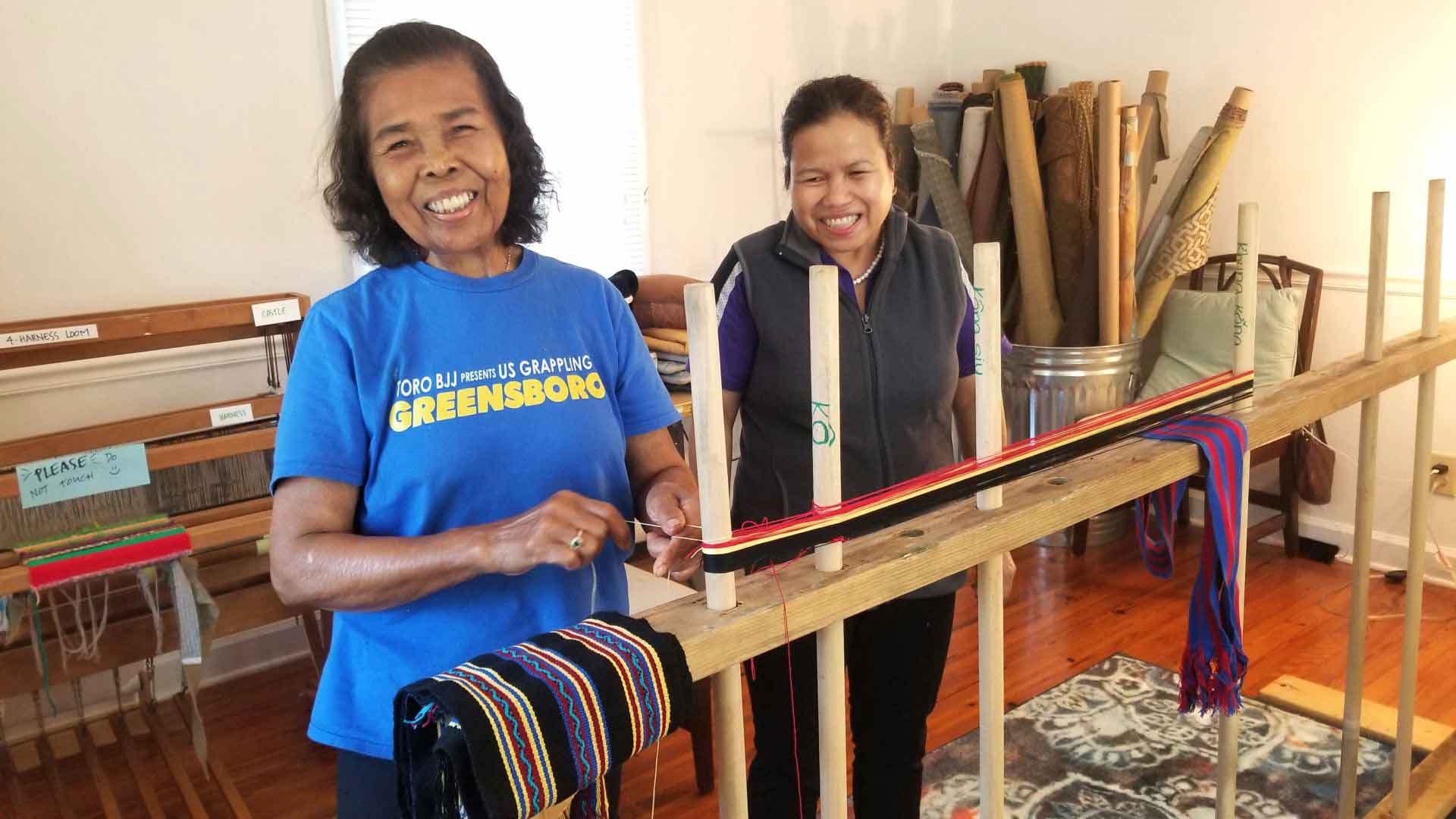 Two older women smile at the camera with a backstrap weaving contraption in front of them.