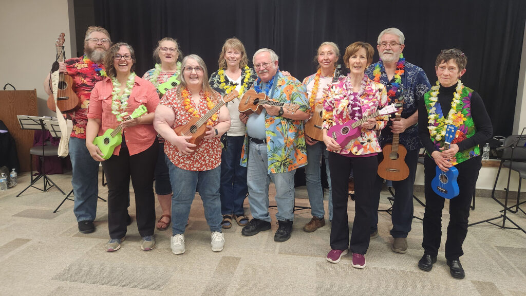 a group of creative aging students from a ukelele class stand together in Hawaiian shirts and leis, holding their instruments and smiling.