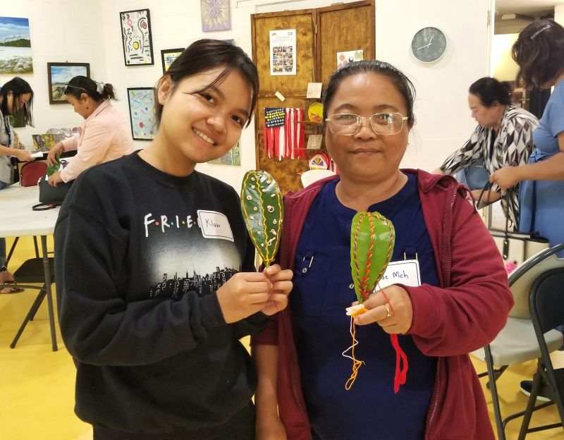 A young teen and older adult pose next to each other holding needlepoint nature artwork they created.