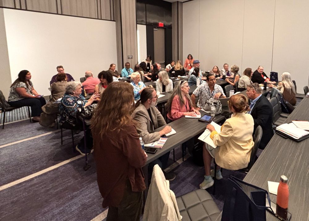 A group of people sit together in a conference room, talking and writing down notes.