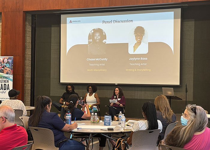 Joylynn sits between two other panelists (Chase McCurdy and Julie Kline) underneath a projector and speaks to an audience sitting around round tables.