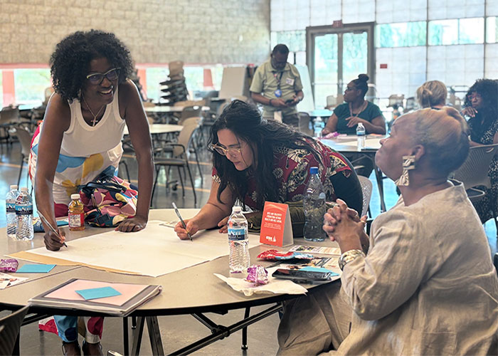 Joylynn (pictured left) leans over a table and writes on a big poster while smiling and looking at two other woman; one is also writing on the poster and the other is sitting in a chair looking at Joylynn.