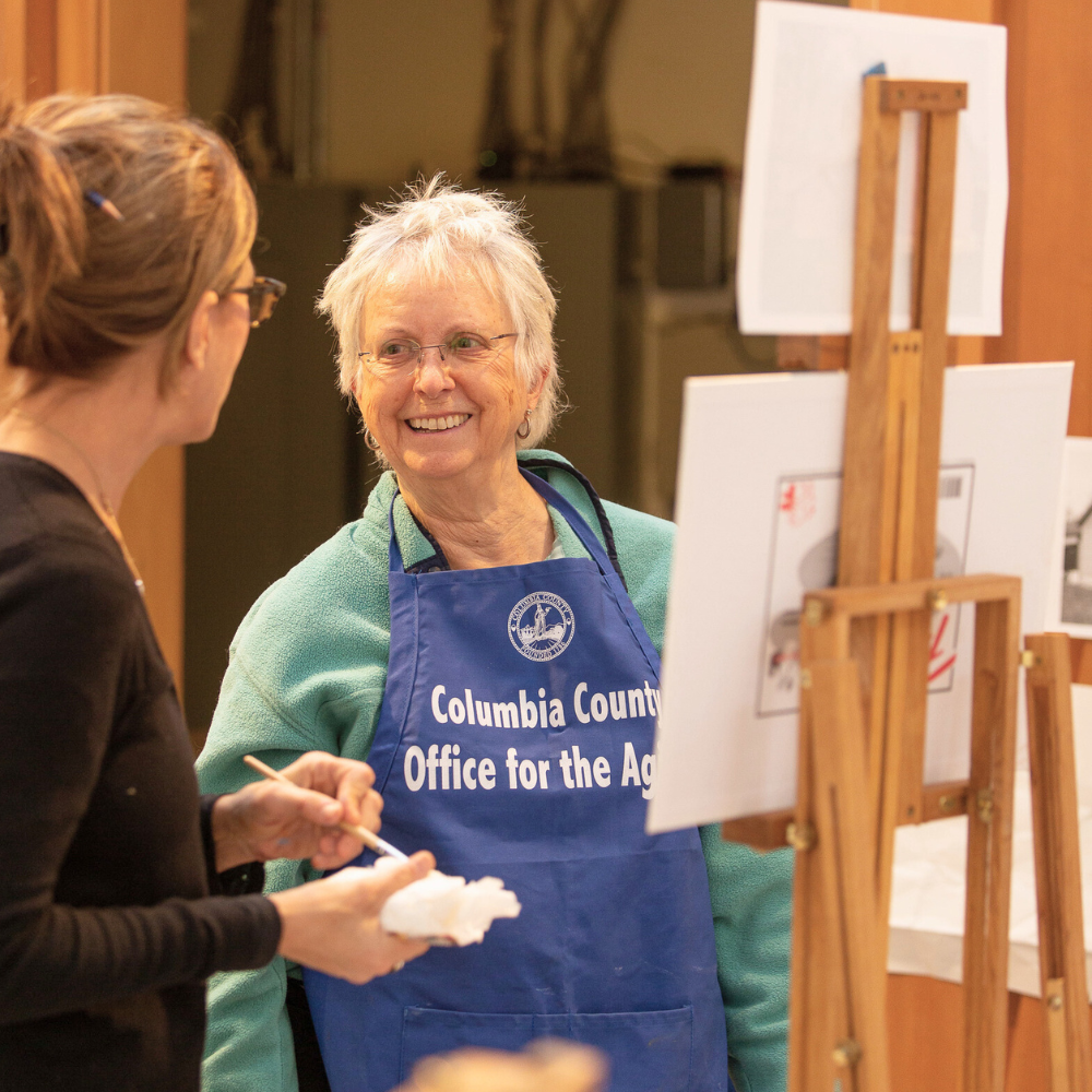 An older woman working at an easel smiles to a teaching artist.