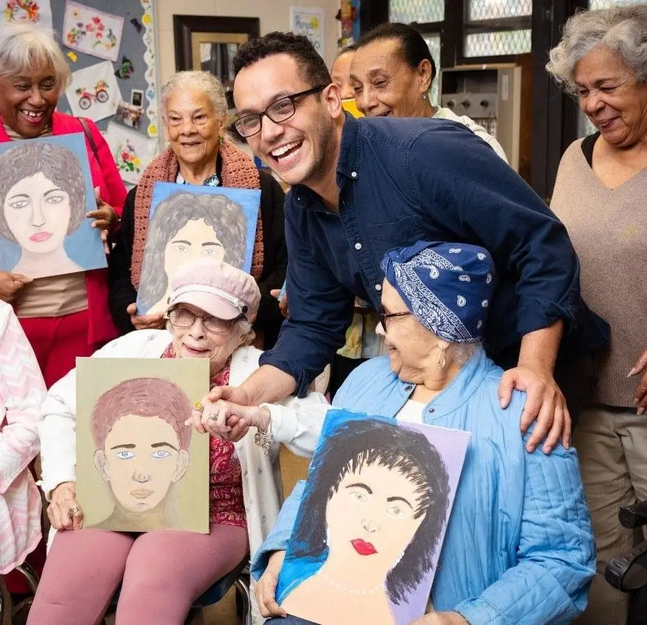 A group of older adult women pose for a group photo holding painted portraits. They're smiling with a younger man.