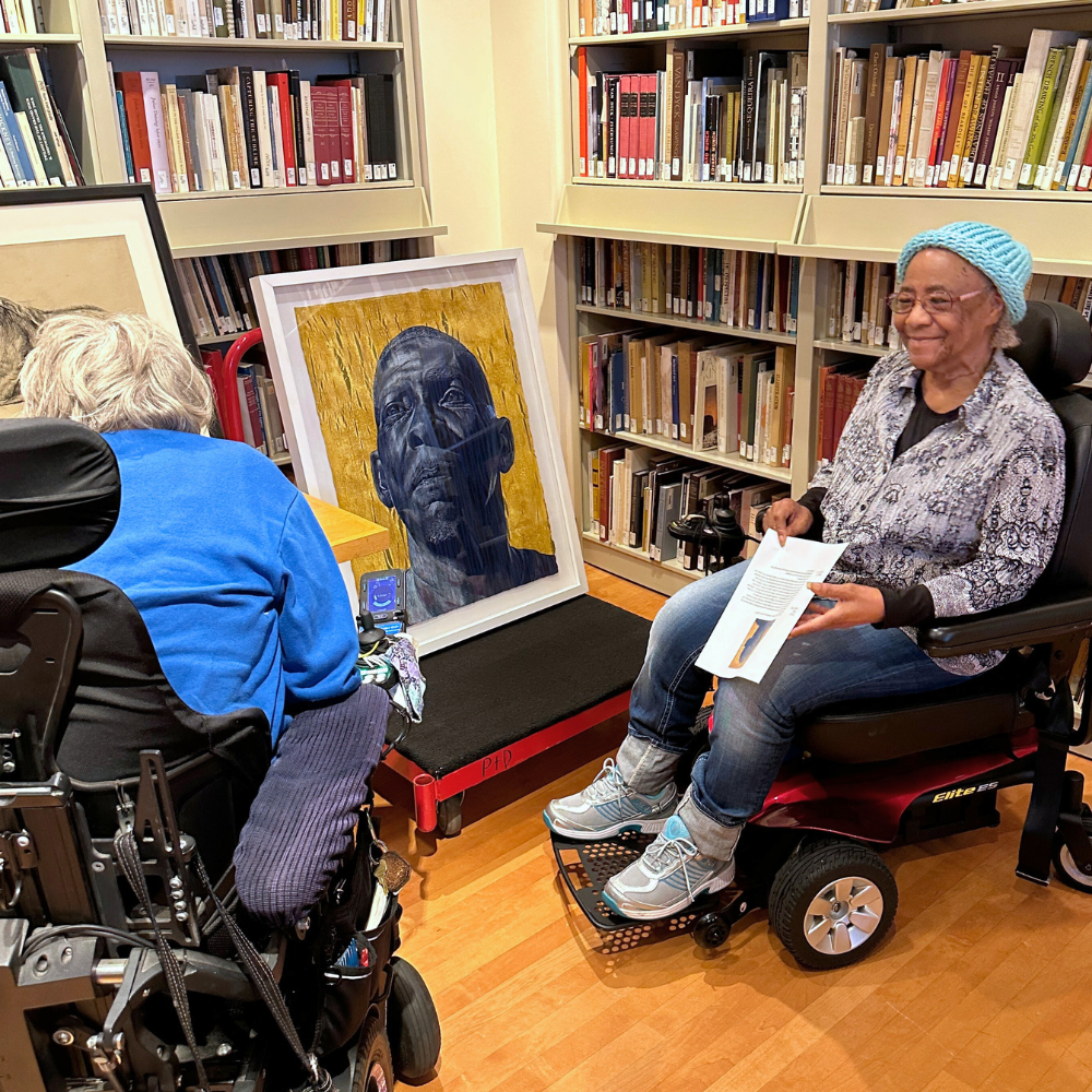 Two older adults in wheelchairs admire artwork surrounded by bookshelves.