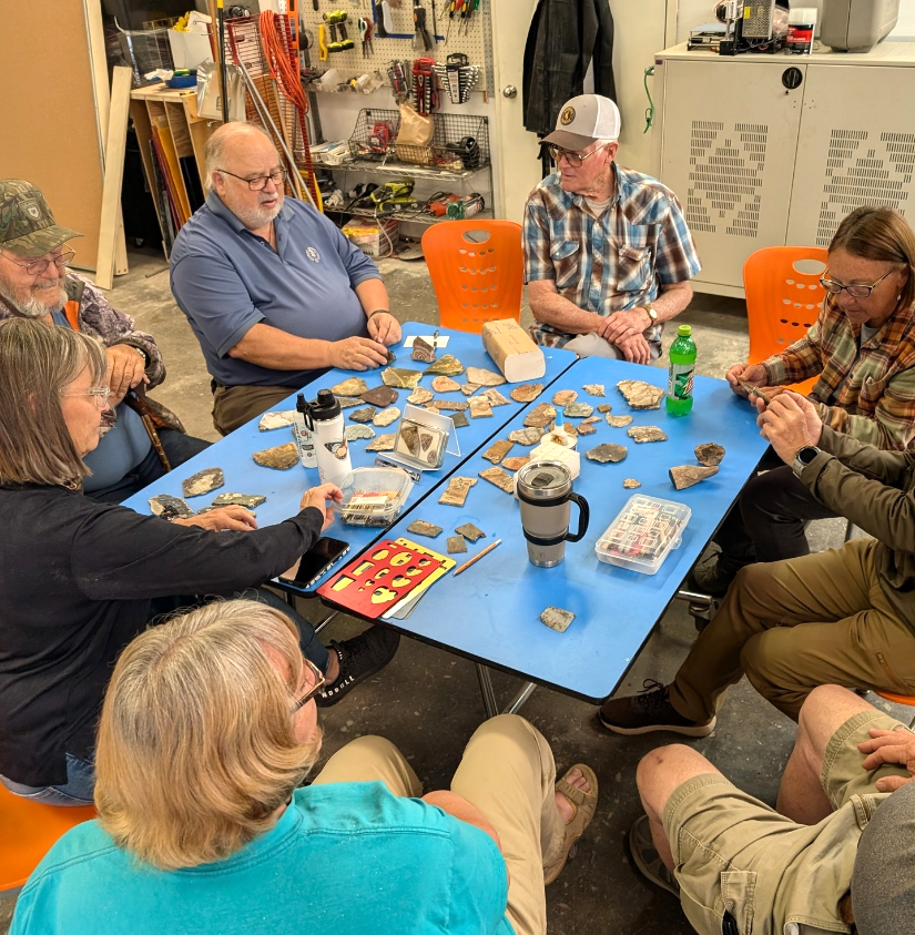 A group of older adults sit around a table together with lapidary materials.
