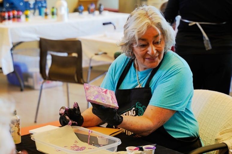 An older adult woman holds a square canvas with paint on it. She is wearing a black apron and is siting at an art table.