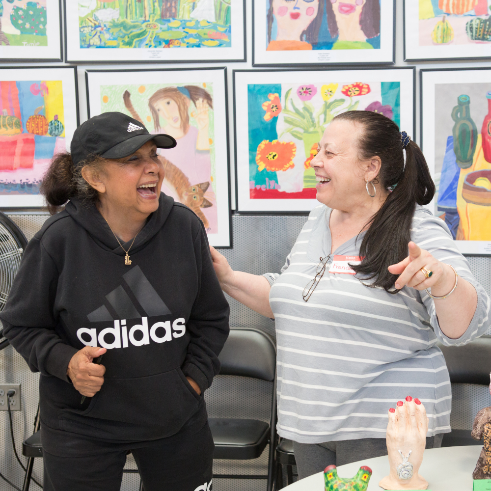 A teaching artist and older woman smile at each other while the teaching artist puts her hand on the older woman's arm.