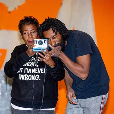 A male teaching artist and older adult female student hold a camera together. The teaching artist looks through the lens.