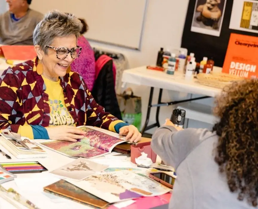 An older woman smiles at a table while flipping through an art magazine.