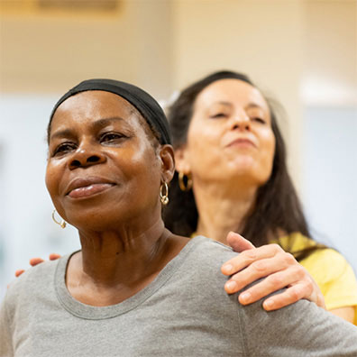 An older Black woman smiles confidently ahead of her. A Latina female teaching artist grabs the older woman's shoulder with her head held high.