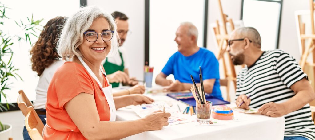 A group of 5 older adults sit around a table painting. one is turned toward the camera smiling.